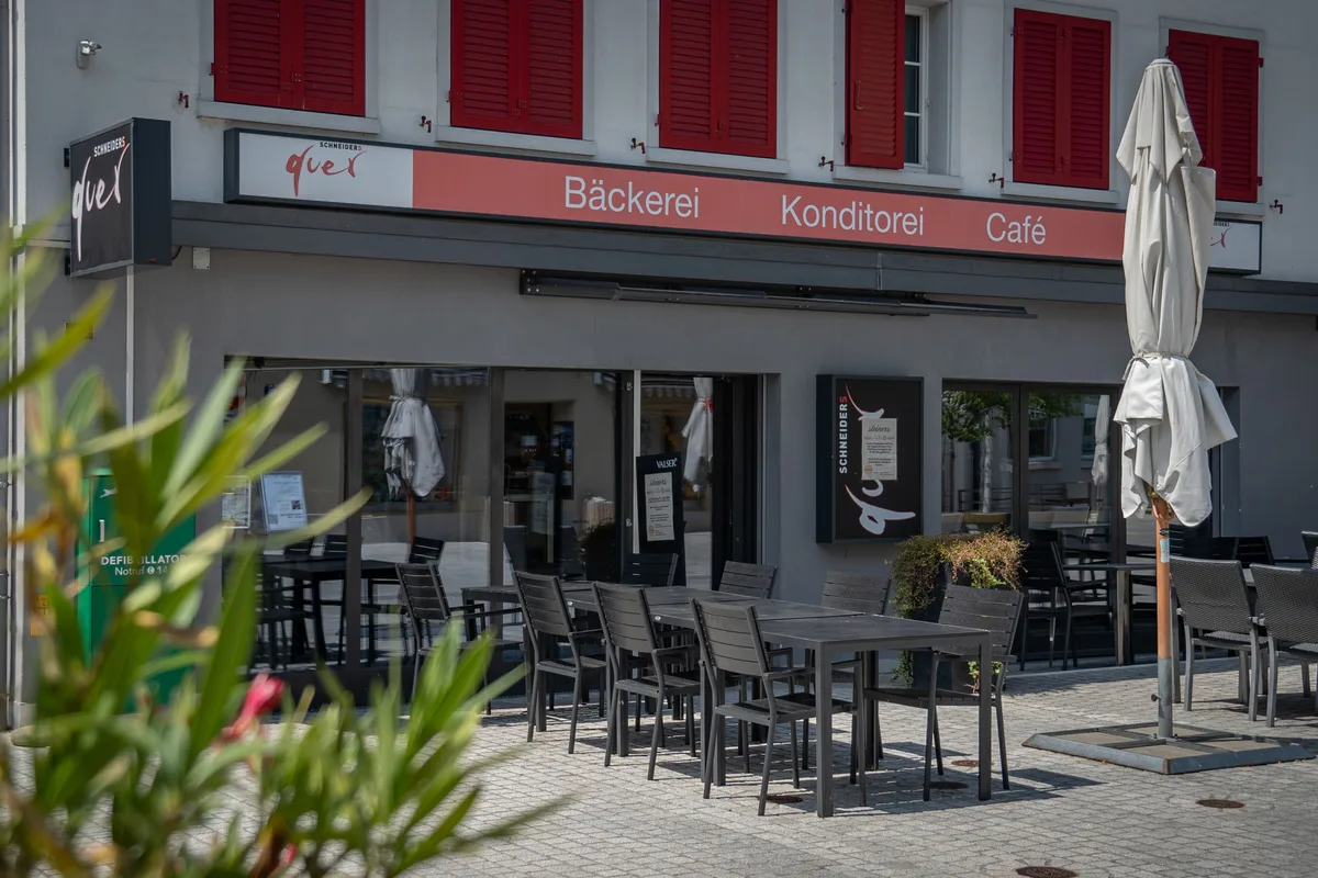Eine kleine Bäckerei-Filiale mit Café in Pfäffikon. Man sieht den Schriftzug «Schneiders Quer – Bäckerei, Konditorei, Café» sowie dunkle Stühle und Tische auf dem Pflaster davor.